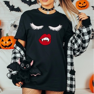 May include: Black t-shirt with a vampire-themed design featuring white eyelashes and red lips with fangs. The shirt is part of a Halloween collection. The person is wearing a black choker and a black and white checkered cardigan.