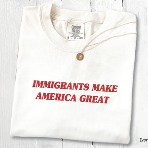 以下が含まれることがあります： 「IMMIGRANTS MAKE AMERICA GREAT」の赤い文字がプリントされたアイボリー色のTシャツ。丸いペンダントが付いたゴールドカラーのネックレスがシャツの上に置かれています。シャツは白い木製の表面に折りたたまれています。
