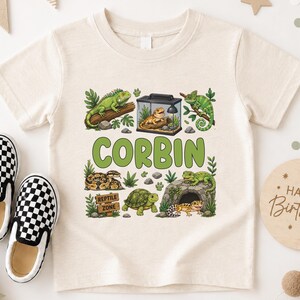 Könnte beinhalten: Ein cremefarbenes Kurzarm-T-Shirt mit einem Grafikdruck verschiedener Reptilien, darunter eine Schlange, eine Schildkröte und Eidechsen, mit dem Namen CORBIN in großen grünen Buchstaben und einem Schild mit der Aufschrift REPTILE ZONE.