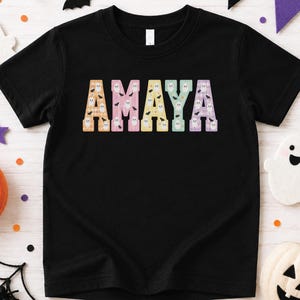 May include: A black short-sleeved t-shirt featuring the name AMAYA printed in large block letters across the chest. Each letter has a unique pastel plaid pattern filled with small white ghost and black bat illustrations.