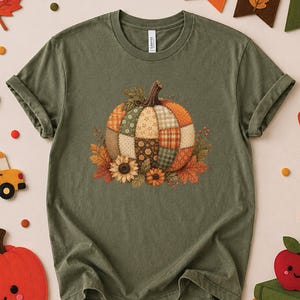 May include: An olive green short-sleeve t-shirt featuring a central graphic of a pumpkin constructed from various patchwork fabric patterns in shades of orange, brown, and green, accented with sunflowers and autumn leaves at the base.