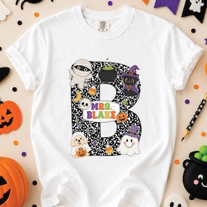 Könnte beinhalten: Ein weißes Kurzarm-T-Shirt mit einem großen schwarz-weißen Notizbuchmuster in Form des Buchstabens B. Der Buchstabe ist mit Halloween-Symbolen wie einer Mumie, einer schwarzen Katze, einem Geist und einem Welpen sowie dem Text MRS. BLAKE verziert.