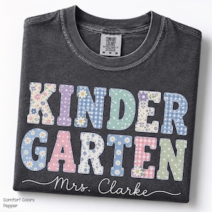 May include: A charcoal gray crew neck t-shirt featuring the word KINDERGARTEN in large, block letters with various pastel floral, gingham, and polka dot patterns. Below the text, the name Mrs. Clarke is printed in a white cursive script font.