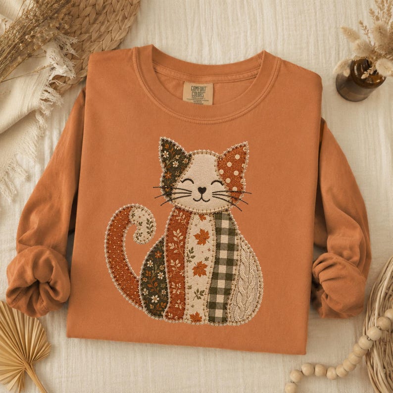 May include: A long-sleeve crewneck sweatshirt in a burnt orange color featuring a central graphic of a cat composed of various fabric-style patches. The patches include patterns of autumn leaves, green and white gingham, and floral prints.