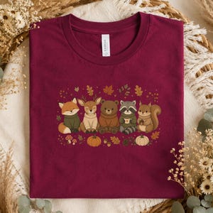 May include: A maroon crew neck t-shirt featuring a graphic print of a fox, deer, bear, raccoon, and squirrel wearing sweaters, surrounded by autumn leaves, mushrooms, and pumpkins. The shirt has a white label at the neck with the text CANVAS.