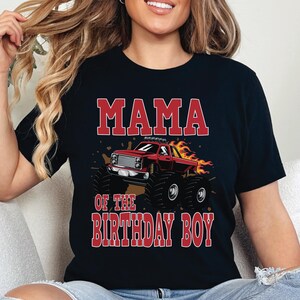 May include: Black t-shirt with the text "MAMA OF THE BIRTHDAY BOY" in red block letters. A red monster truck with flames is featured in the center. The shirt is a crew neck style.