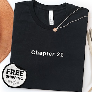 Peut inclure: T-shirt noir à col rond avec le texte "Chapter 21" en blanc. Un collier argenté avec un pendentif circulaire repose sur le t-shirt. Un chapeau marron et des feuilles décoratives sont visibles en arrière-plan.