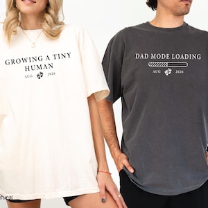 May include: Two matching t-shirts, one ivory and one charcoal. The ivory shirt reads "GROWING A TINY HUMAN" with "AUG 2026" below. The charcoal shirt says "DAD MODE LOADING" with a loading bar and "AUG 2026".