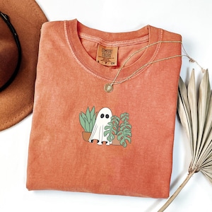 Puede incluir: Una camiseta naranja Comfort Colors con un diseño de fantasma de dibujos animados y plantas en macetas. El fantasma es blanco con ojos negros y las plantas son verdes. Un collar dorado con un colgante está sobre la camisa.