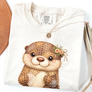 以下が含まれることがあります： クリーム色のTシャツ。可愛いラッコのイラストがハートを持っています。ラッコはパッチワーク生地で作られ、花の冠をかぶっています。金のネックレスと茶色の帽子も写っています。