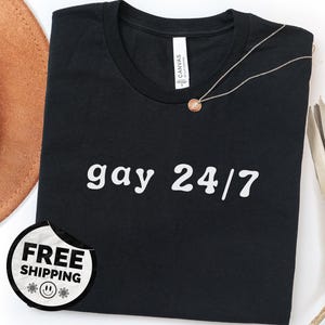 May include: Black t-shirt with the phrase "gay 24/7" in white lettering. A silver necklace with a small pendant rests on the shirt. A brown hat and dried plants are visible in the background.