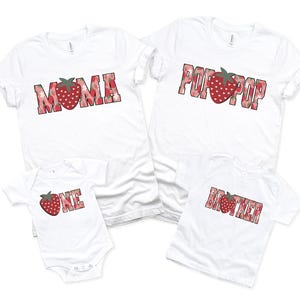Puede incluir: Dos camisetas blancas de manga corta, un body blanco para beb&eacute; y una camiseta blanca para ni&ntilde;o peque&ntilde;o. Las prendas presentan letras de bloque con estampado de fresas rojas que deletrean MOMA, POP POP, ONE y BROTHER, con una fresa sustituyendo la O.