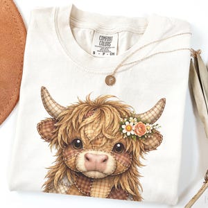Puede incluir: Camiseta Comfort Colors color crema con una ilustración de una vaca de las Tierras Altas con una corona de flores. La vaca tiene pelaje y cuernos de estilo patchwork. Un collar dorado con un colgante descansa sobre la camiseta.