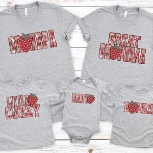 Puede incluir: Una colecci&oacute;n de cinco prendas de color gris jaspeado que incluye tres camisetas y un body para beb&eacute;. Cada pieza presenta un texto con estampado de fresas rojas que dice GRANDPA, GREAT GRANDMA, LITTLE CUZZY, BABY y BUBBA, con fresas rojas.