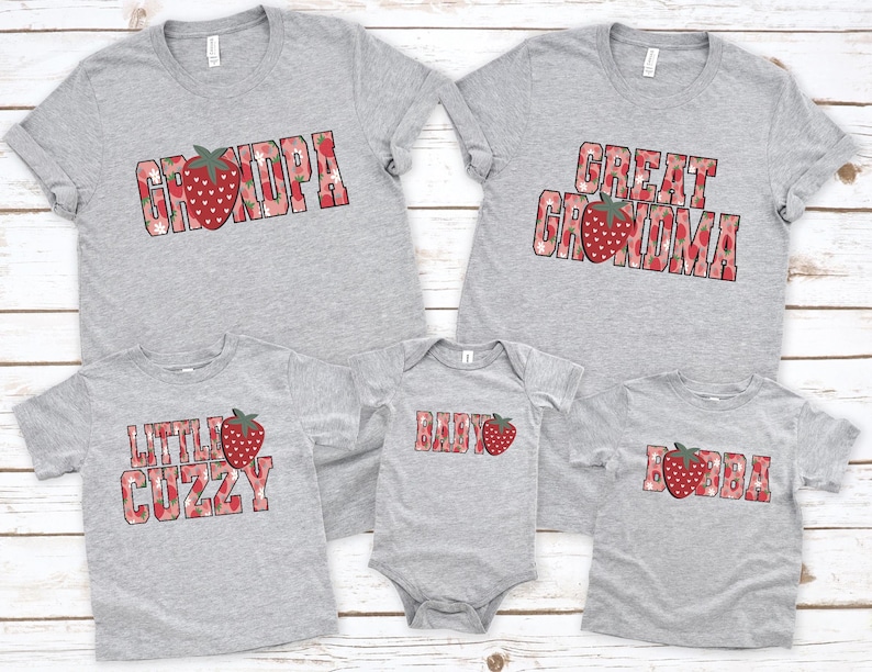 Puede incluir: Una colecci&oacute;n de cinco prendas de color gris jaspeado que incluye tres camisetas y un body para beb&eacute;. Cada pieza presenta un texto con estampado de fresas rojas que dice GRANDPA, GREAT GRANDMA, LITTLE CUZZY, BABY y BUBBA, con fresas rojas.