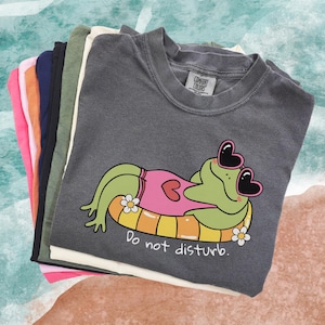 Może przedstawiać: Stos koszulek Comfort Colors w różnych kolorach, w tym szarym, różowym i granatowym. Górna koszulka jest szara i przedstawia kreskówkową żabę w okularach przeciwsłonecznych w kształcie serca i różowym kostiumie kąpielowym, wylegiwującą się na żółto-pomarańczowej dmuchanej zabawce. Pod żabą znajduje się napis "Do not disturb".