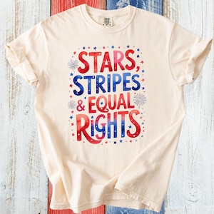 Op de afbeelding: Crèmekleurig T-shirt met de tekst "STARS, STRIPES & EQUAL RIGHTS" in rood en blauw, omringd door sterren en vuurwerk. Het shirt wordt getoond op een houten oppervlak met rood, wit en blauw gestreepte planken.