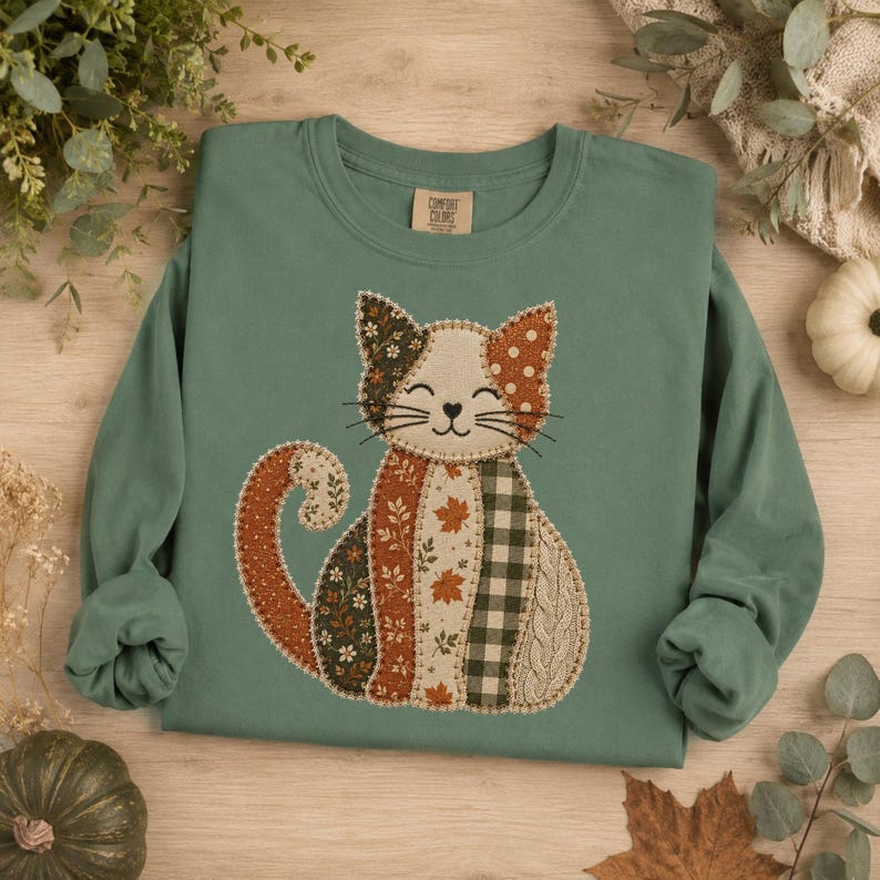 May include: A sage green long-sleeve sweatshirt featuring a central graphic of a cat composed of various fabric-style patches. The cat design includes patterns of autumn leaves, dark green plaid, polka dots, and cable knit textures in earth tones.