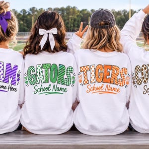 Puede incluir: Cuatro sudaderas blancas de cuello redondo con gráficos en la espalda que muestran nombres de equipos en varios patrones y colores. El texto dice TEAM Your Team Name Here, GATORS School Name, TIGERS School Name y COWBOYS School Name en morado, verde, naranja y marrón.