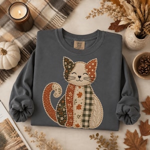 Peut inclure: Un sweat-shirt à col rond gris anthracite orné d'un motif central de chat composé de divers motifs de style tissu, notamment des feuilles d'automne, du vichy vert et des imprimés floraux. Le chat a un contour cousu et un petit nez en cœur.