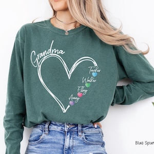 May include: A teal-colored sweatshirt with the word "Grandma" in cursive script above a white heart design. Inside the heart are the names "Tucker", "Walker", "Elley", and "Avery", each with a colored heart. The text "Blue Spurce" is at the bottom right.