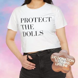 Puede incluir: Top corto blanco con el texto "PROTECT THE DOLLS" en negro. La modelo lleva vaqueros negros. Una pegatina con forma de flor con el texto "$10 DONATION TO TRANS LIFELINE" es visible.