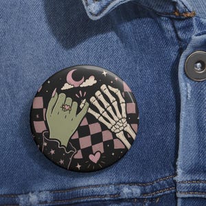 May include: A circular pin button featuring a graphic illustration of a green hand and a skeletal hand performing a pinky promise. The background displays a black and dusty pink checkered pattern with small stars, a crescent moon, and a heart.