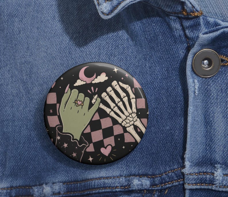 May include: A circular pin button featuring a graphic illustration of a green hand and a skeletal hand performing a pinky promise. The background displays a black and dusty pink checkered pattern with small stars, a crescent moon, and a heart.