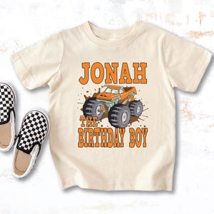 May include: Cream-colored toddler t-shirt featuring the name "Jonah" and the phrase "The Birthday Boy" in orange block letters. A cartoon monster truck in orange and teal is centered below the name. Two black and white checkered slip-on shoes are to the left.