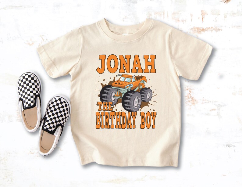 May include: Cream-colored toddler t-shirt featuring the name "Jonah" and the phrase "The Birthday Boy" in orange block letters. A cartoon monster truck in orange and teal is centered below the name. Two black and white checkered slip-on shoes are to the left.