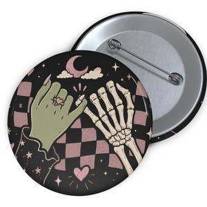 May include: A round pin-back button featuring a graphic illustration of a green hand and a skeleton hand performing a pinky promise. The background displays a black and dusty pink checkered pattern with small stars, a crescent moon, and a heart.