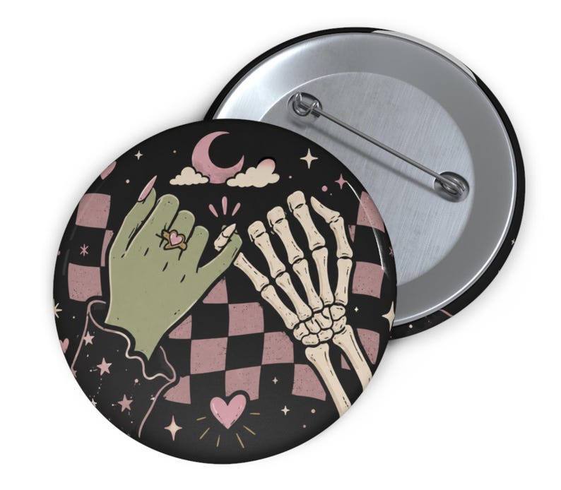 May include: A round pin-back button featuring a graphic illustration of a green hand and a skeleton hand performing a pinky promise. The background displays a black and dusty pink checkered pattern with small stars, a crescent moon, and a heart.
