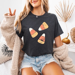 May include: Dark gray t-shirt featuring three candy corn graphics. The candy corn design includes white, yellow, and orange colors. The shirt is a short-sleeved crew neck.