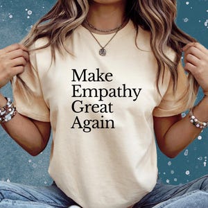 Puede incluir: Camiseta color crema con el texto negro "Make Empathy Great Again". La camiseta está siendo sostenida por una persona. La persona lleva pulseras y collares.