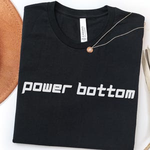 May include: Black t-shirt with the words "power bottom" in white, block letters. A delicate silver necklace with a small, round pendant rests on the shirt. A brown hat and dried plant are visible in the background.