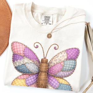 May include: Cream-colored t-shirt featuring a patchwork butterfly design with pink, purple, blue, and yellow wings. The butterfly has a brown body and antennae. A gold necklace with a pendant rests on the shirt. A brown hat and dried palm leaf are also visible.