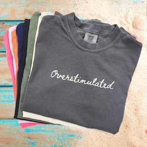May include: A stack of t-shirts in various colors, including pink, orange, green, and black. The top shirt is dark gray with the text "Overstimulated" in white.
