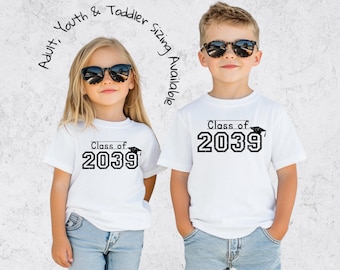 May include: Two children wearing white t-shirts with the text "Class of 2039" in black, along with a graduation cap graphic. The shirts are paired with blue jeans. The text "Adult, Youth & Toddler sizing Available" is arched above the children.