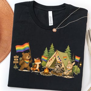May include: Black t-shirt featuring a camping scene with cartoon animals, rainbow flags, and a tent. The design includes a bear, fox, raccoon, and rabbit enjoying a campfire. The text "Love is Love" is on a sign.