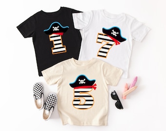 May include: Three white, black and beige t-shirts with a pirate hat and number design. The shirts feature the numbers 1, 5 and 7 with black and white stripes. The pirate hats are blue and red with a skull and crossbones design.