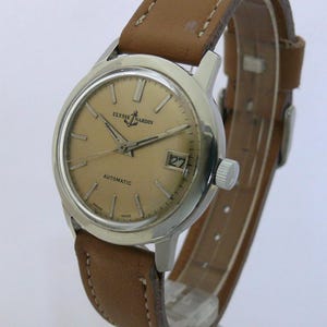 May include: A vintage Ulysse Nardin automatic wristwatch with a light brown leather strap. The watch face is a light gold colour with silver hands and markers. The date is displayed at the 3 o'clock position. The watch has a silver case.