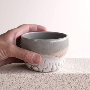 Puede incluir: Un pequeño cuenco de cerámica gris con un esmalte blanco y un borde texturizado. El cuenco se sostiene en una mano.