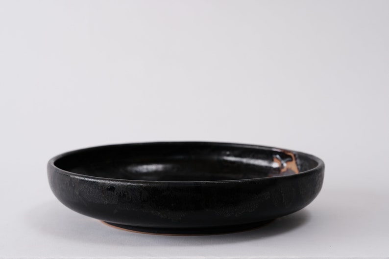 stunning handmade stoneware low serving bowl with a soft contemporary curve leading to the elevating foot ring. The bowl is finished in a metallic textured black glaze with a ray of unglazed part in the center.