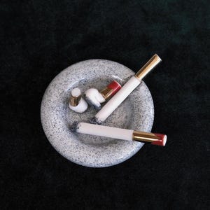 May include: A speckled gray ashtray with several white and gold-tipped cigarettes and cigarette butts. The cigarettes have red tips. The ashtray is round and has a speckled pattern. The background is a dark green.