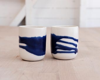 May include: Two cylindrical ceramic cups with a smooth white finish, each featuring a distinct hand-painted deep blue abstract design. The cup on the left displays a wide horizontal blue stroke, while the cup on the right shows vertical blue lines.