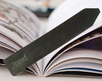 May include: A dark olive green rectangular leather bookmark with a pointed end. The name Josef is embossed in a serif font on the lower portion of the bookmark. The bookmark is placed diagonally across the open pages of a white paper book.