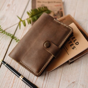 May include: A brown leather notebook cover with a snap button closure and visible stitching, containing a Field Notes memo book. A black and gold pen lies nearby. The cover features a simple rectangular design with rounded corners and a strap.