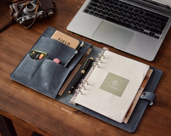 May include: A blue leather planner cover featuring internal pockets holding a tan Field notebook, a black pen, and a small pink eraser. The open planner contains lined paper with a square green sticker displaying the text ts and calligraphy.