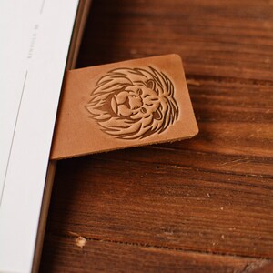 May include: A rectangular tan leather bookmark featuring a debossed lion head design with a detailed mane. The bookmark is inserted into a white book, with the lion motif clearly visible on the smooth, light brown surface of the leather material.