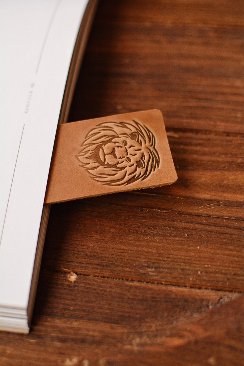 May include: A rectangular tan leather bookmark featuring a debossed lion head design with a detailed mane. The bookmark is inserted into a white book, with the lion motif clearly visible on the smooth, light brown surface of the leather material.